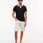 A man modeling a black short-sleeve polo shirt with white trim on the collar and sleeve cuffs, paired with off-white shorts and dark brown boat shoes, against a plain white studio background.