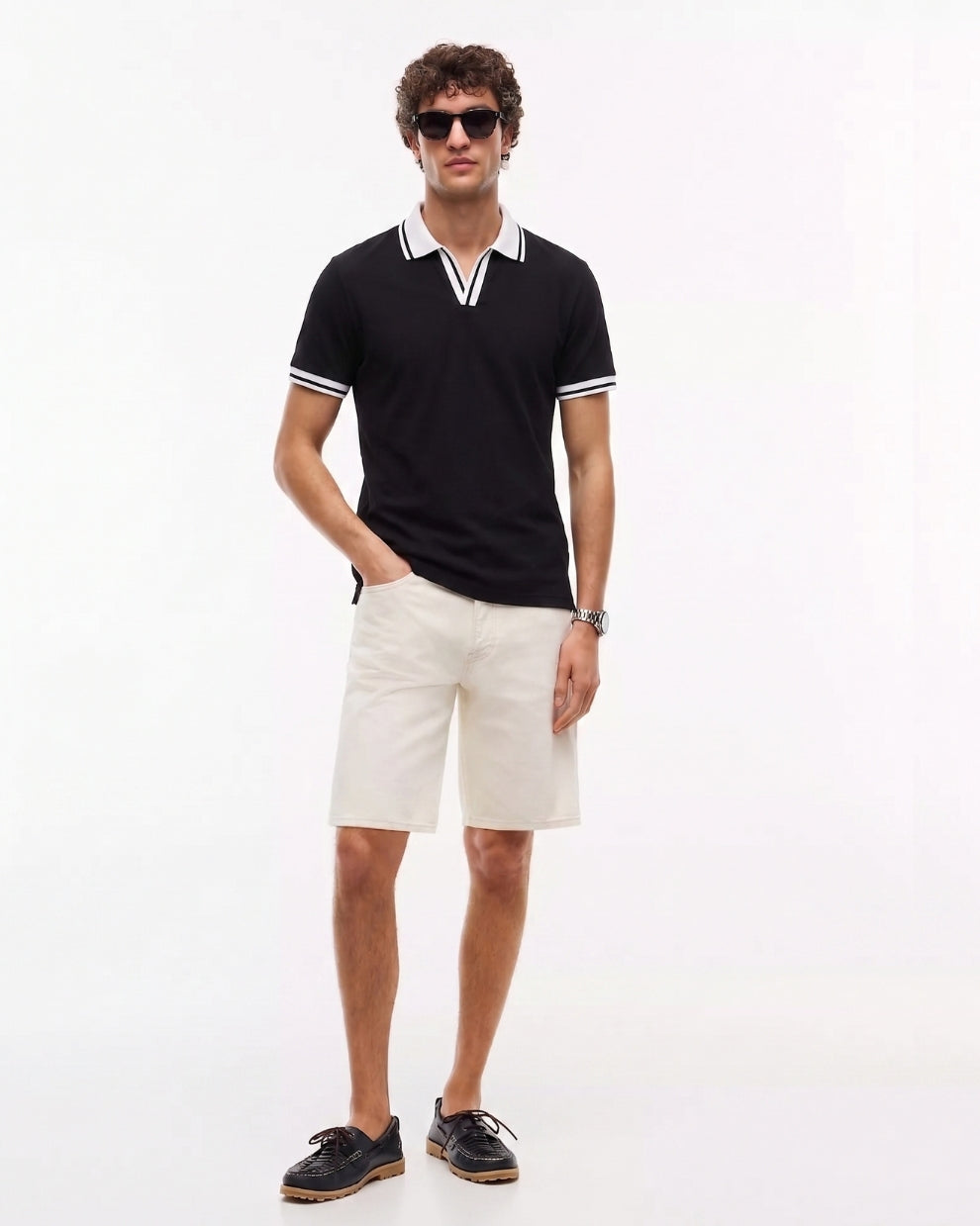 A man modeling a black short-sleeve polo shirt with white trim on the collar and sleeve cuffs, paired with off-white shorts and dark brown boat shoes, against a plain white studio background.